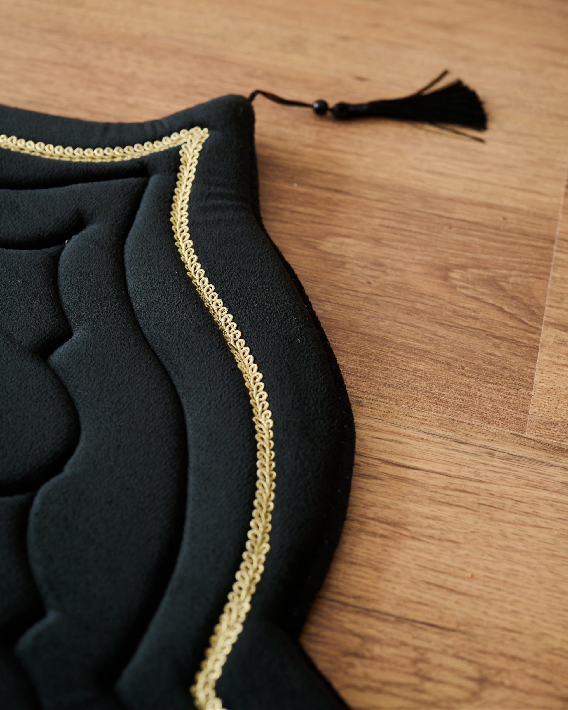 Black memory foam padded prayer mat on a wooden floor with a white sofa and decorative plants in the background
