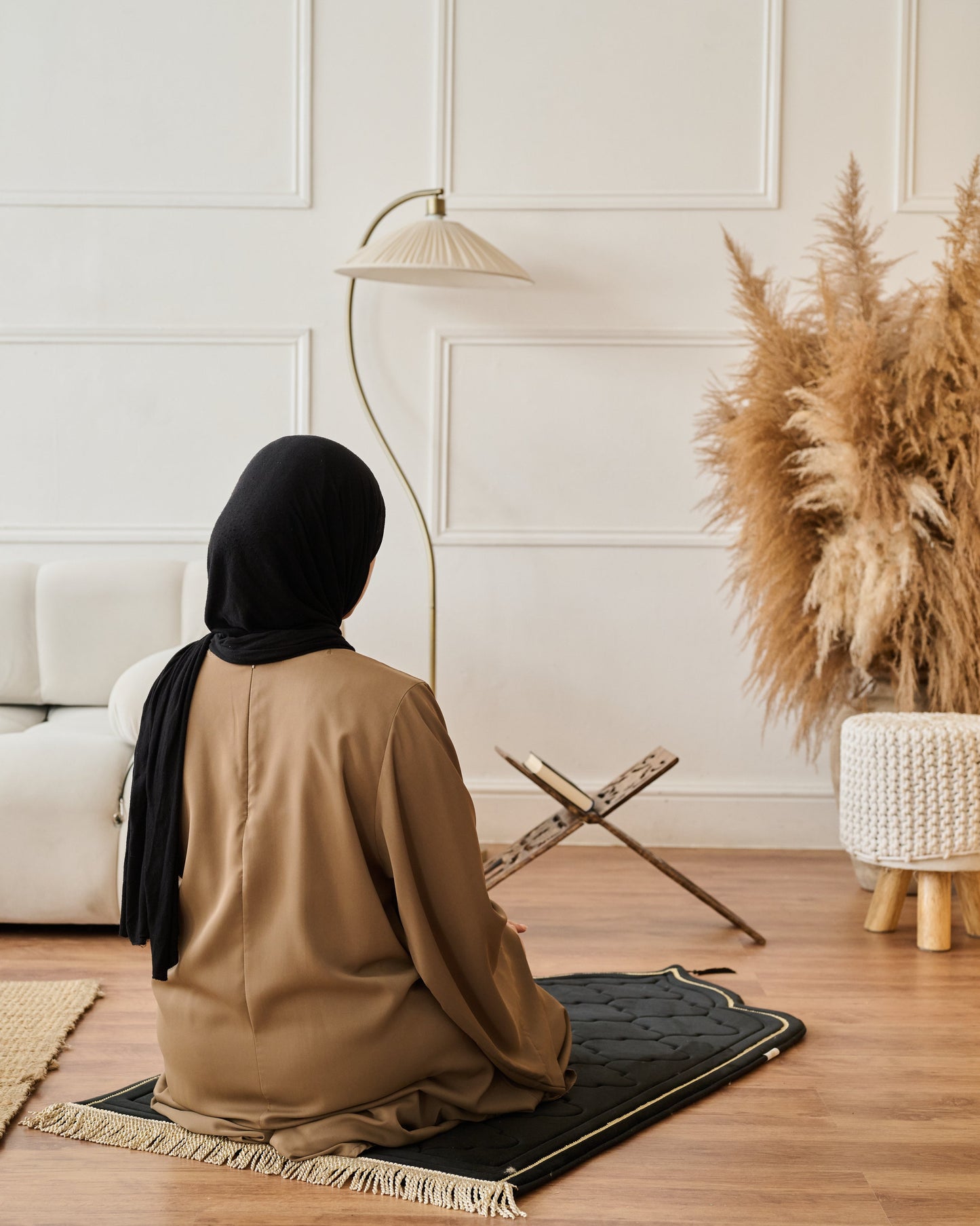 Person in a brown outfit and black headscarf sitting on a prayer mat in a room with a lamp and decorative plant.