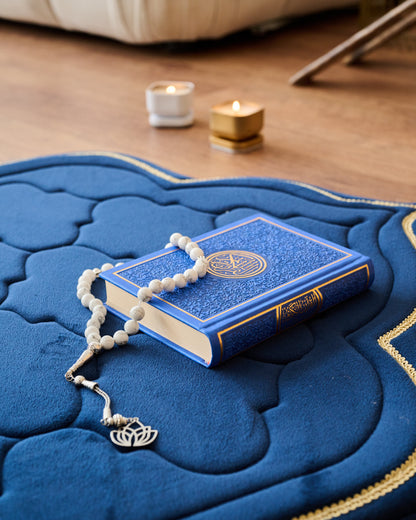 Blue prayer rug with a Quran and rosary on a wooden floor.