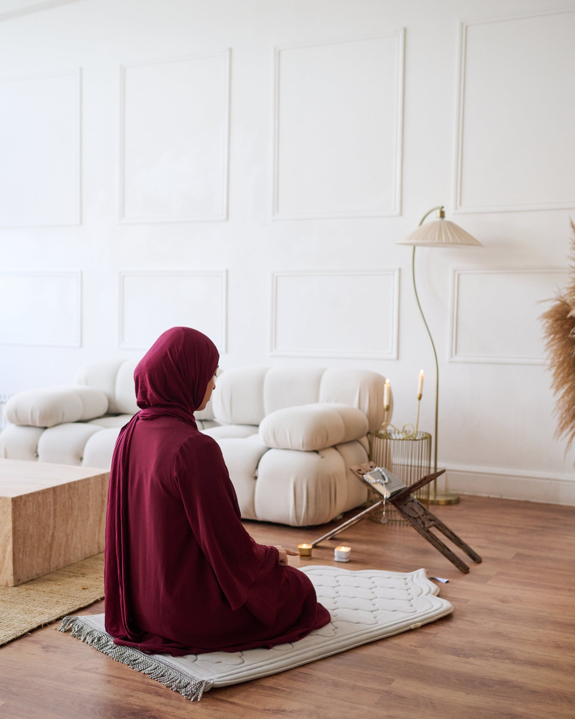 Person in a burgundy hijab and long garment sitting on a prayer mat in a modern living room.