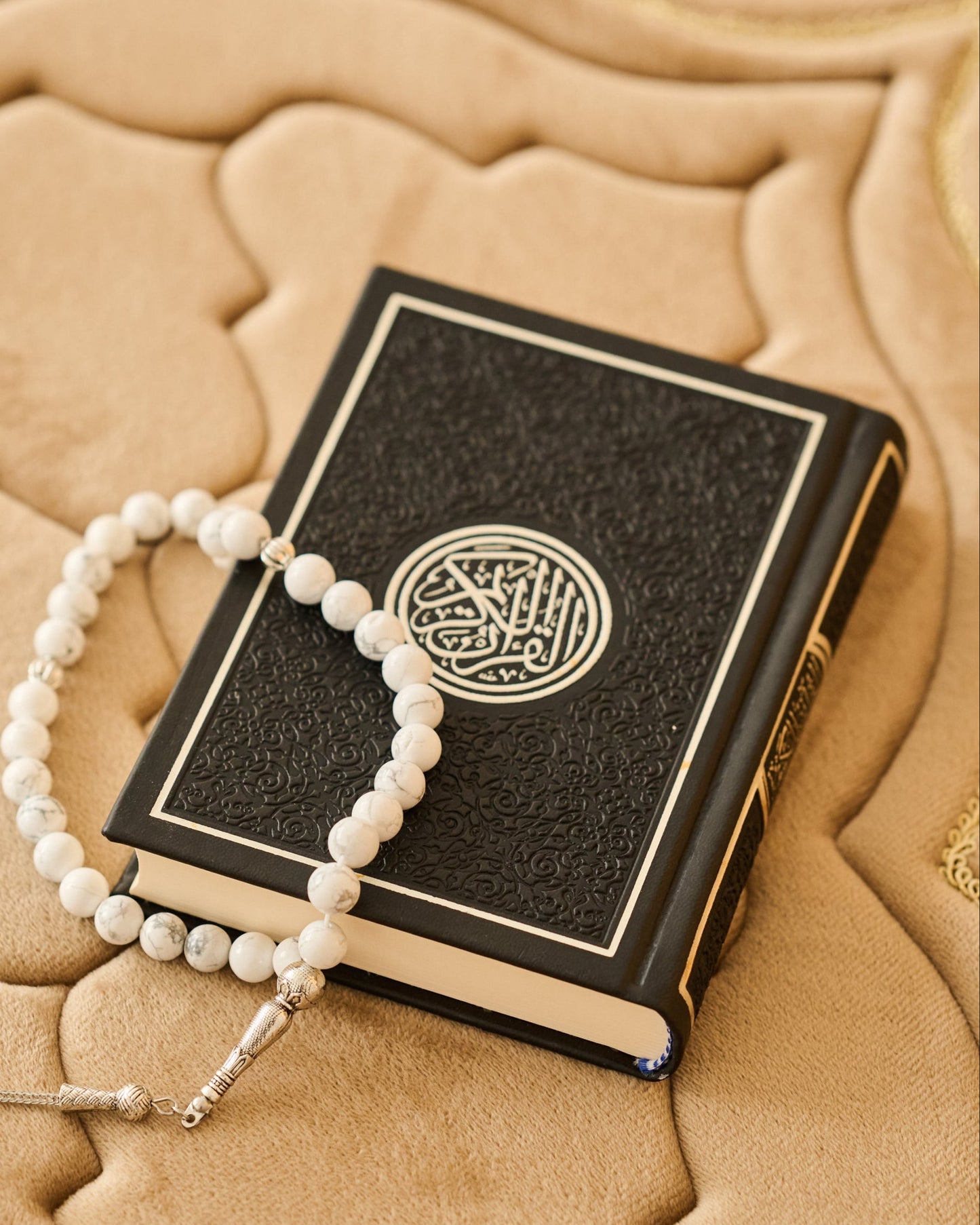 Black book with gold design and white prayer beads on a textured beige surface